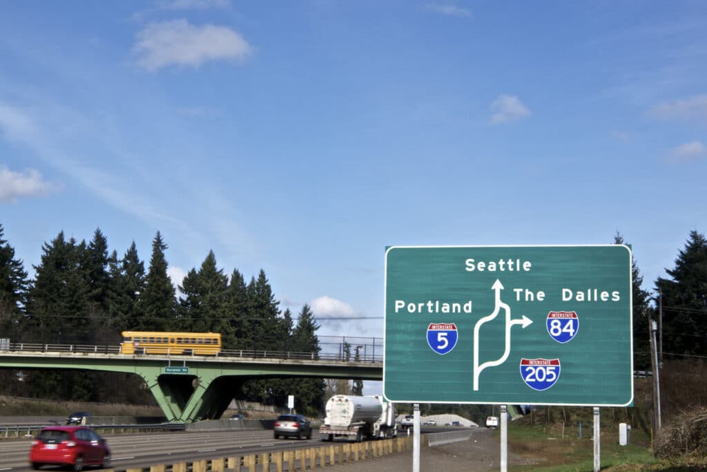 A Large green sign shows direction to Portland, OR , Seattle, WA and The Dalles, OR on Interstate 5 ( I-5 ). A Drawing shows the layout of the roads. Motion blurred traffic on the freeway includes cars, a truck and a school bus. This is a major bypass / alternate route near Portland, Oregon.